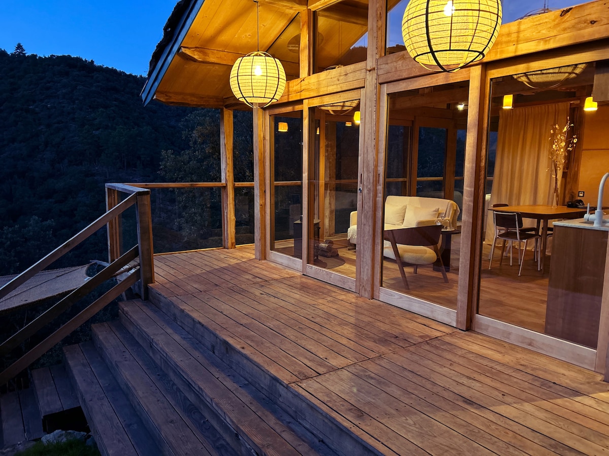 A spacious wooden terrace extends from the lodge, surrounded by nature. Large glass doors invite natural light, while two decorative lanterns hang overhead. The seating area features a soft white couch, offering a relaxing spot to enjoy the serene mountain views.