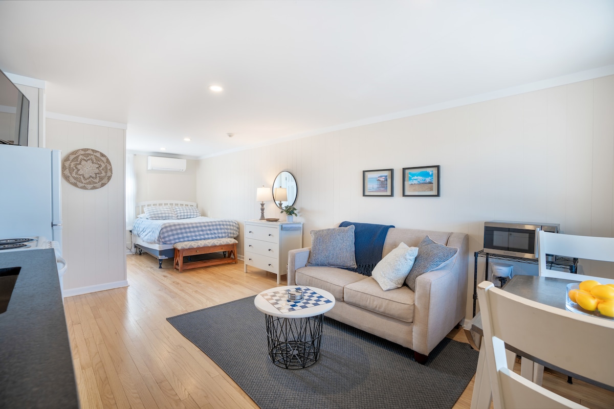 An open studio space features a comfortable full bed against a light-colored wall. A cozy seating area with a sofa and decorative pillows is arranged beside a small, round coffee table. A kitchenette is visible, along with a dining table set for two, enhancing the functional layout.