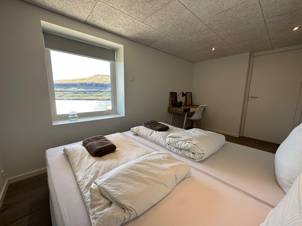 A restful sleeping area is presented with two spacious beds, topped with neatly folded towels. A window provides a scenic view of the surrounding landscape, allowing natural light to illuminate the room. A desk and chair are positioned nearby, offering a functional workspace.