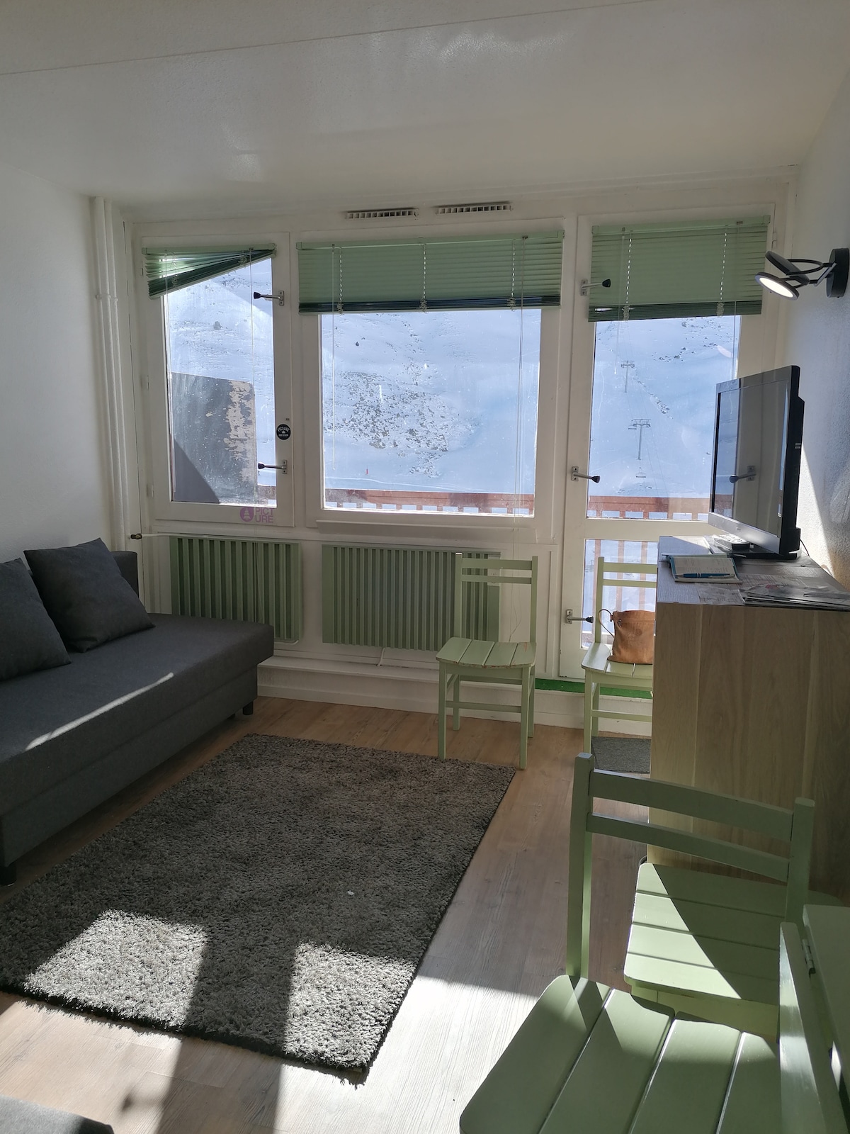 A living area features a comfortable gray sofa paired with a small green table. Natural light pours in through large windows, offering a view of the outside. A flat-screen TV is mounted on a light wood unit, and a soft rug covers the floor.