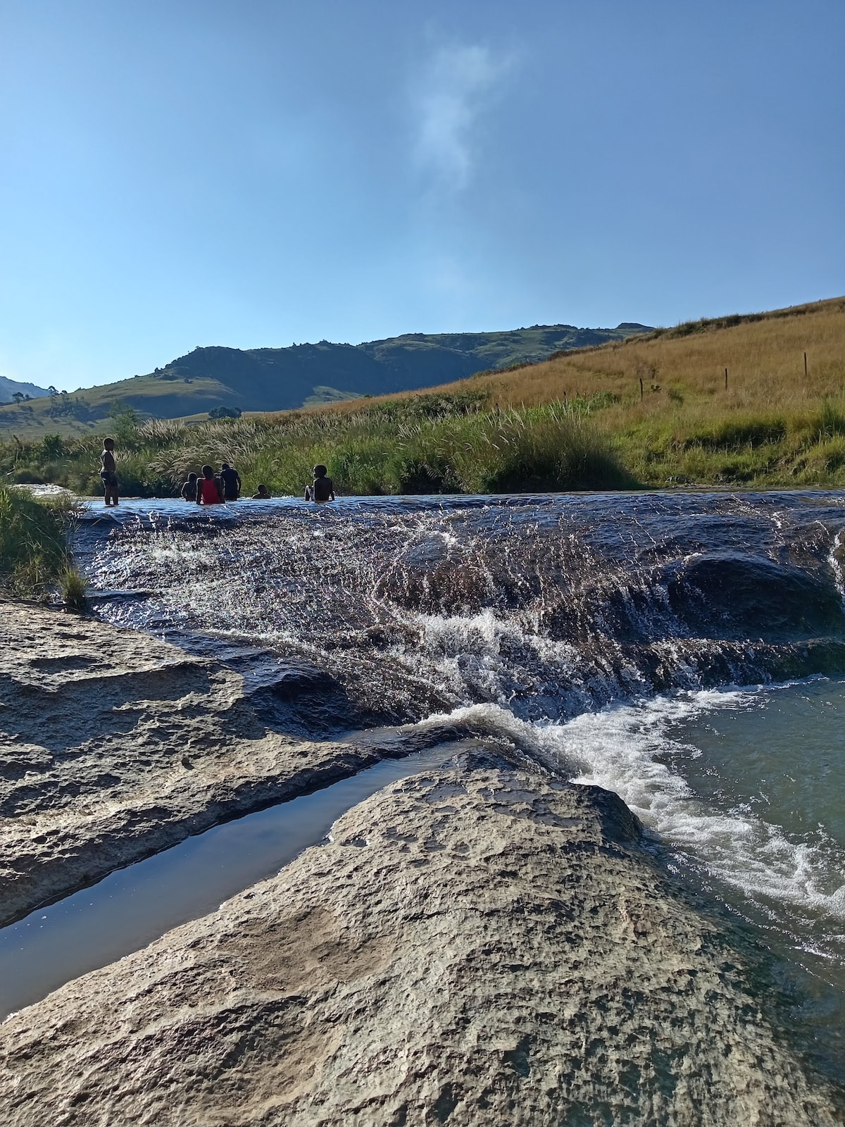 Gentle river waves flow over smooth rocks, with a backdrop of rolling hills and clear blue skies. A few individuals can be seen in the shallow waters, enjoying the natural setting surrounded by lush greenery and open fields.