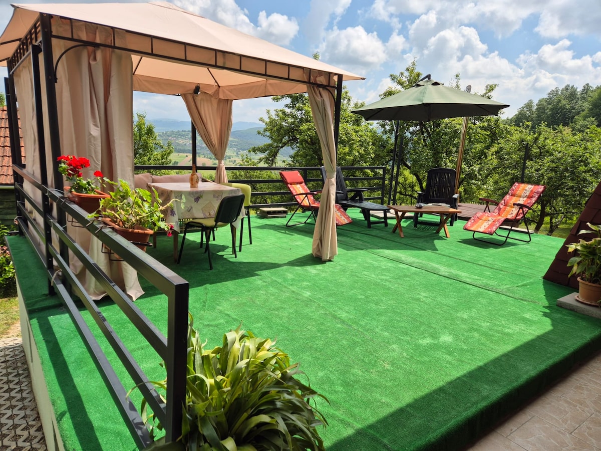 An outdoor terrace is presented, featuring a shaded gazebo with a table and chairs. Sunbeds are arranged for relaxation on a green carpeted surface, alongside a large umbrella providing additional shade. Lush greenery and scenic views enhance the tranquil setting.
