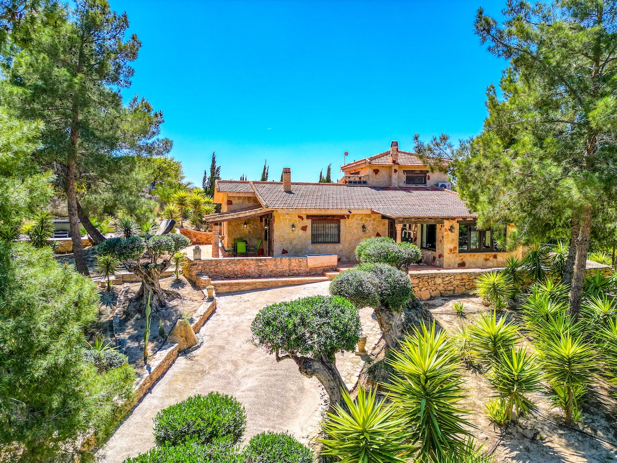 A spacious exterior view presents a rustic stone house surrounded by lush greenery and well-maintained gardens. Pathways meander through the landscape, offering a connection to the natural environment. The clear blue sky complements the warm tones of the building, creating an inviting setting.