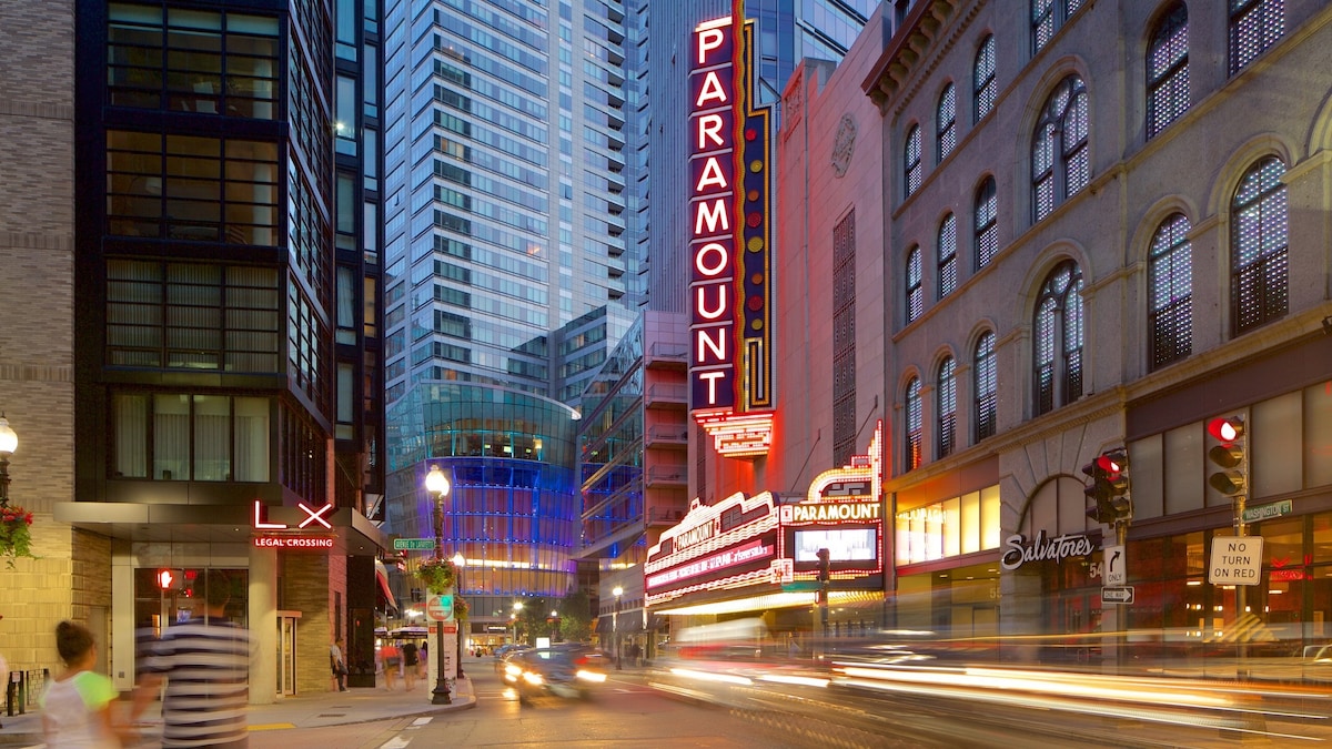 A vibrant urban scene captures the illuminated Paramount Theatre, showcasing its iconic marquee. Tall modern buildings frame the street, accentuated by glowing signs. Pedestrians walk along the crosswalk, while vehicles pass, adding to the bustling atmosphere of downtown Boston.