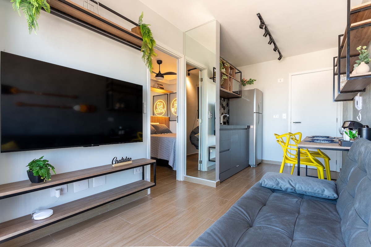 A modern living space is displayed with a large flat-screen television mounted on a wooden shelf. A gray sofa faces the TV, while a bright yellow dining chair is set near a small desk. A mirrored cabinet reflects light, enhancing the room's openness.