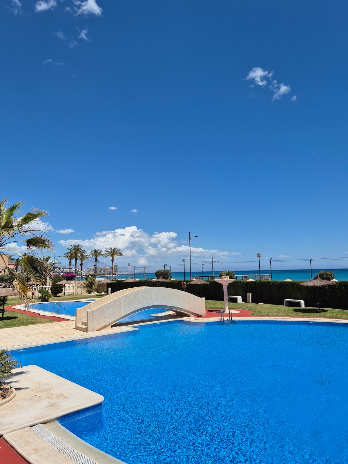 Two inviting swimming pools are set against a clear blue sky, with swimming areas reflecting sunlight. Lush palm trees surround the pools, and a bridge arches over the water, leading to spacious areas for relaxation. The Mediterranean coastline is visible in the background.