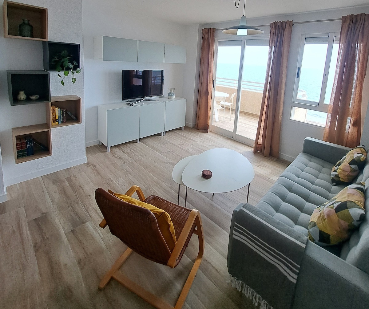 A living area is displayed, featuring a comfortable gray sofa with decorative pillows, a circular coffee table, and a woven armchair. Large glass doors lead to a terrace with panoramic sea views. Light curtains frame the windows, allowing natural light to fill the space.