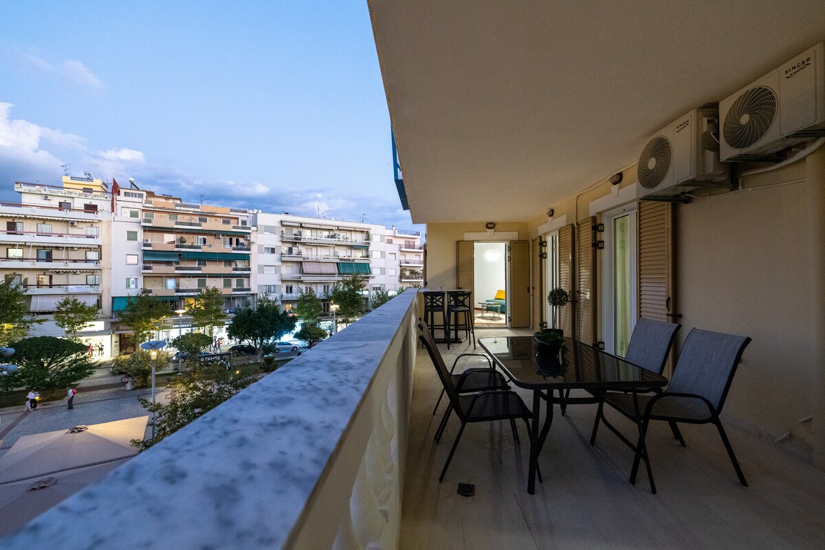 A spacious balcony features a table and four chairs, ideal for relaxation. The view includes buildings and trees, with soft lighting enhancing the evening atmosphere. Air conditioning units are visible on the side wall, contributing to comfort in warm weather.