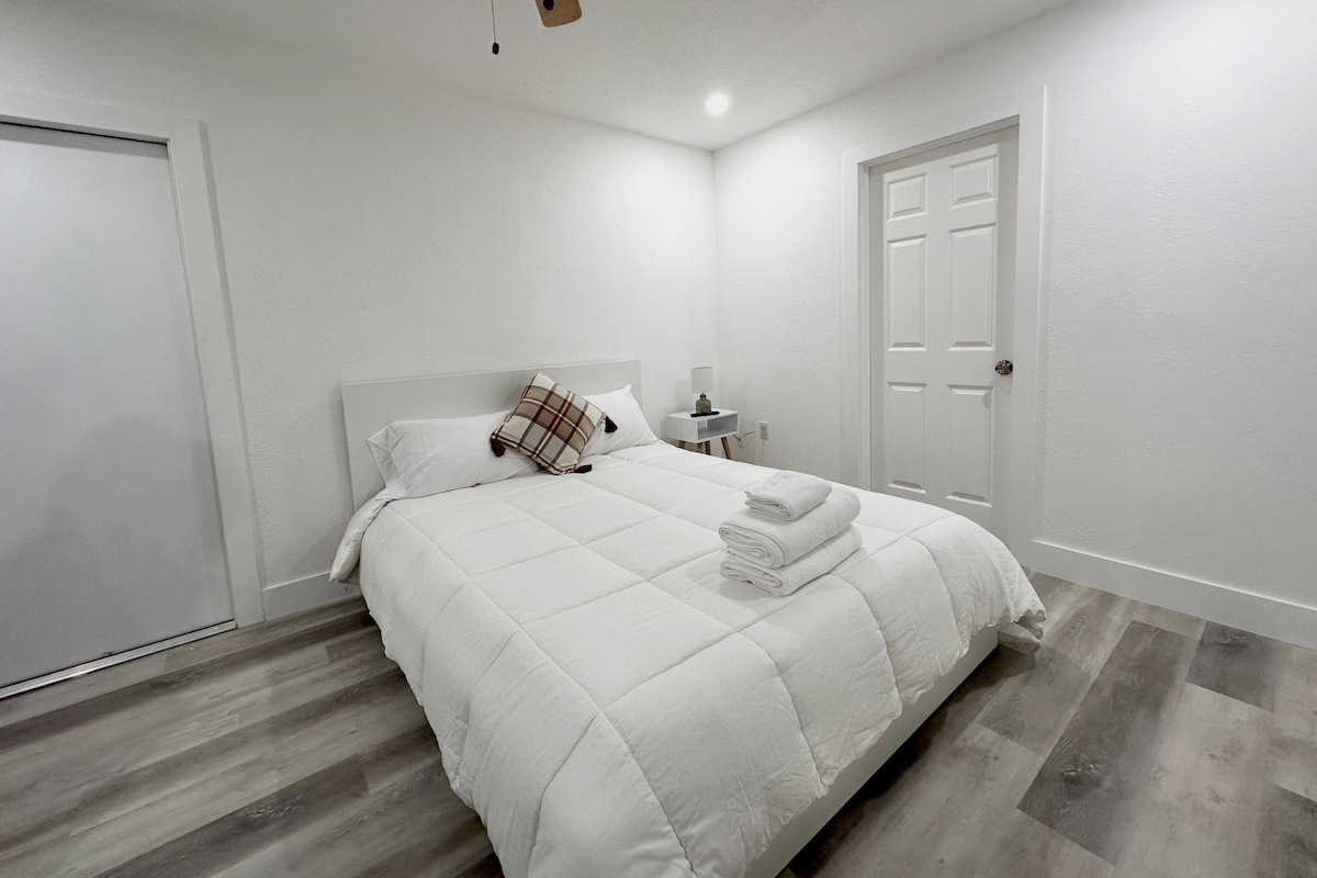 A well-lit bedroom features a spacious bed with a crisp white comforter and decorative pillows. White walls and neutral flooring create a calming environment. A simple nightstand and a closed closet are visible, contributing to the room's functional layout.
