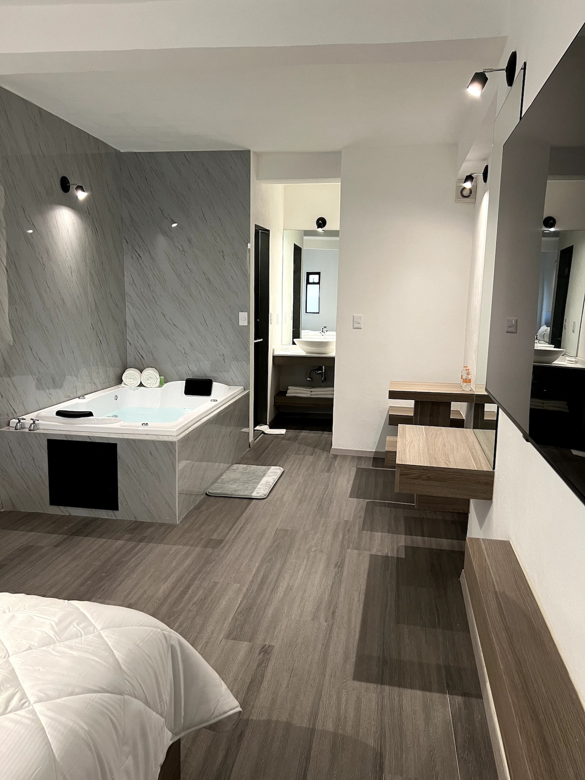 A spacious suite features a bathroom with a Jacuzzi tub surrounded by elegant marble accents. A cozy sleeping area is visible, with a large flat-screen smart TV mounted on the wall. Light-colored wooden furniture enhances the modern aesthetic.