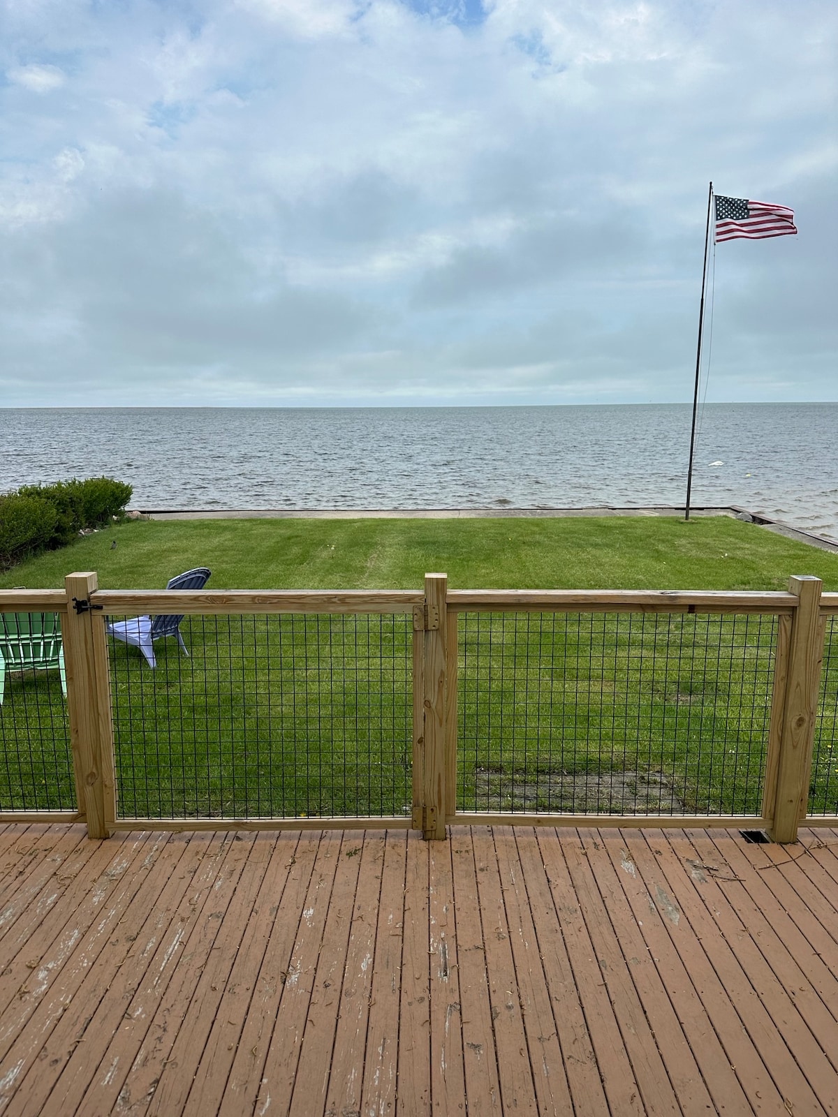 A grassy area extends towards Saginaw Bay, framed by a wooden deck. An American flag is proudly displayed on a pole, with a single blue chair visible on the lawn. Calm water stretches across the horizon, under a partly cloudy sky.