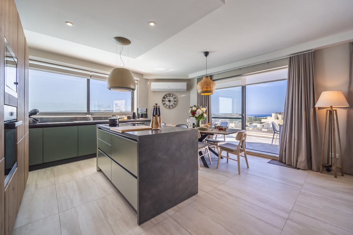 A modern kitchen with sleek cabinetry and an island counter is presented. Large windows provide ample natural light and offer views of the outdoor terrace. A dining area features a wooden table surrounded by chairs, while stylish light fixtures add to the contemporary design.