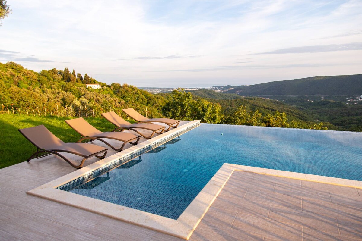 An infinity pool is set against a scenic landscape, featuring a gentle gradient from light blue to deep blue water. Four lounge chairs are positioned along the pool's edge, facing towards the expansive views of the hills and valleys under a clear sky.