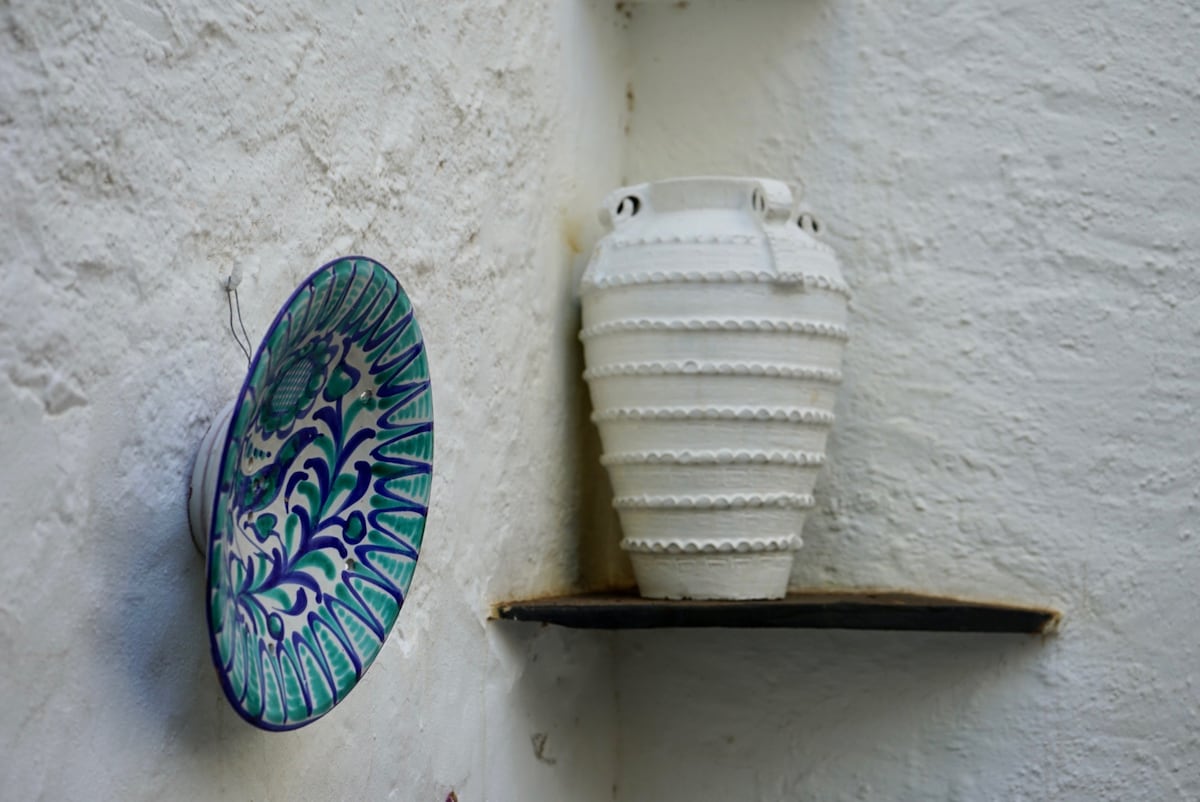 A decorative plate with intricate blue and green patterns is displayed on the wall beside a textured white vase. The vase has a unique ribbed design and is positioned on a simple black shelf, enhancing the visual appeal of the cream-colored wall.