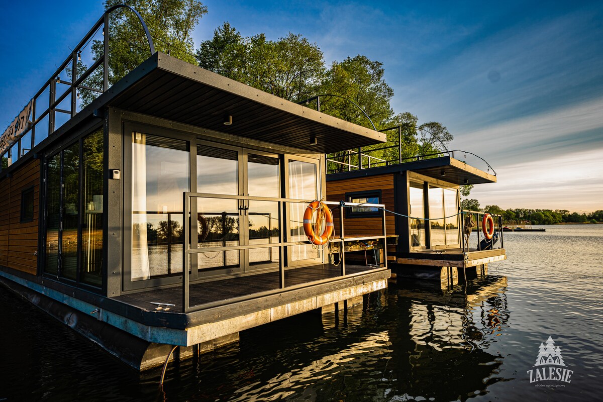 Two modern floating cabins are positioned on calm waters, featuring expansive glass doors that reflect the surrounding greenery. Each cabin has a spacious deck with a safety ring and railings, enhancing the seamless connection between indoor and outdoor relaxation.