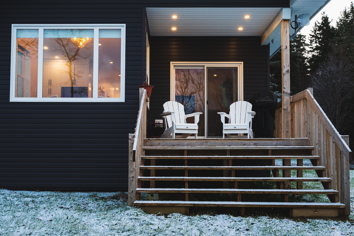 A wooden deck features two white, adirondack-style chairs facing outward. The entrance is illuminated by soft lighting, highlighting the dark exterior of the house. Snow covers the ground, creating a serene atmosphere.