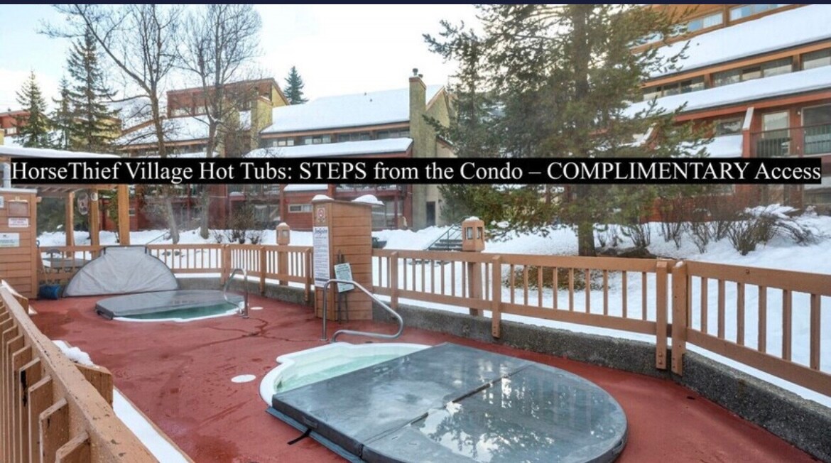 The image captures the outdoor hot tub area at Horsethief Village, featuring two hot tubs surrounded by wooden fencing. Snow blankets the ground, while the nearby buildings provide a cozy backdrop. Steps lead from the condo directly to this inviting relaxation space.