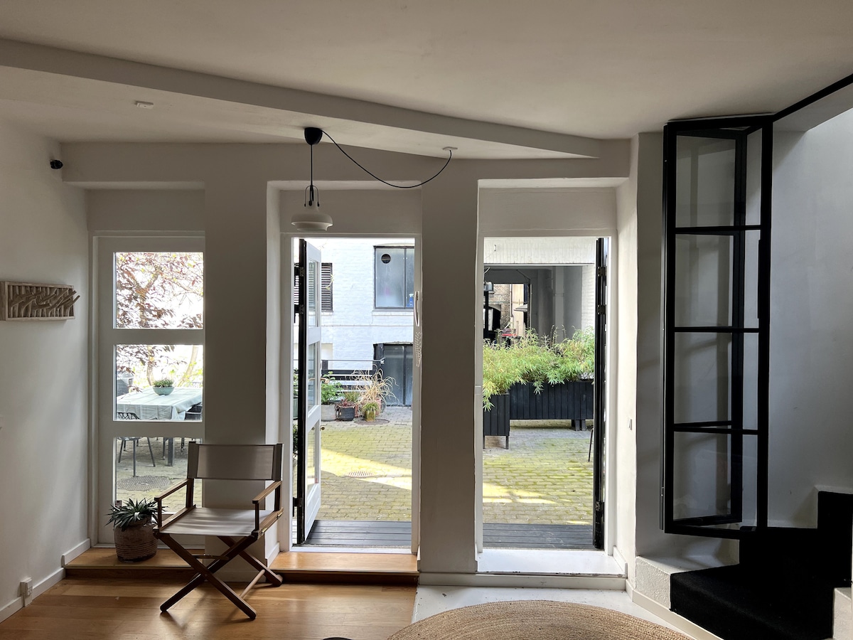Two glass doors open to a serene courtyard, allowing natural light to filter into the spacious living area. A stylish chair and a small plant add subtle detail, while the contrasting black spiral staircase leads to the upper levels of the townhouse.