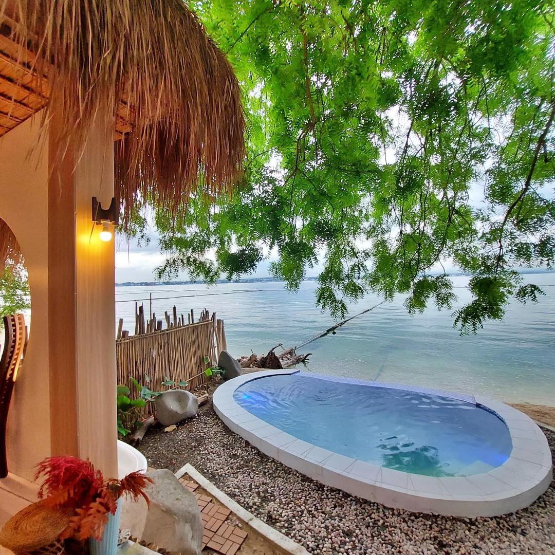 A small but inviting pool is situated beside the shoreline, surrounded by smooth stones and gentle waves. Green foliage provides shaded areas, and a thatched roof is visible in the background, enhancing the connection with nature.