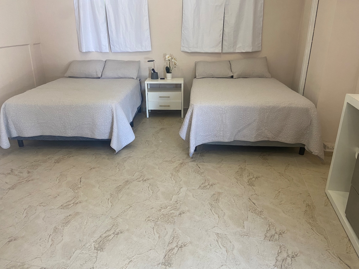 The sleeping area features two spacious beds draped with light-colored bedspreads. A nightstand is positioned between them, complemented by a small decorative flower arrangement. Natural light filters in through two windows adorned with sheer curtains, enhancing the calm ambiance.