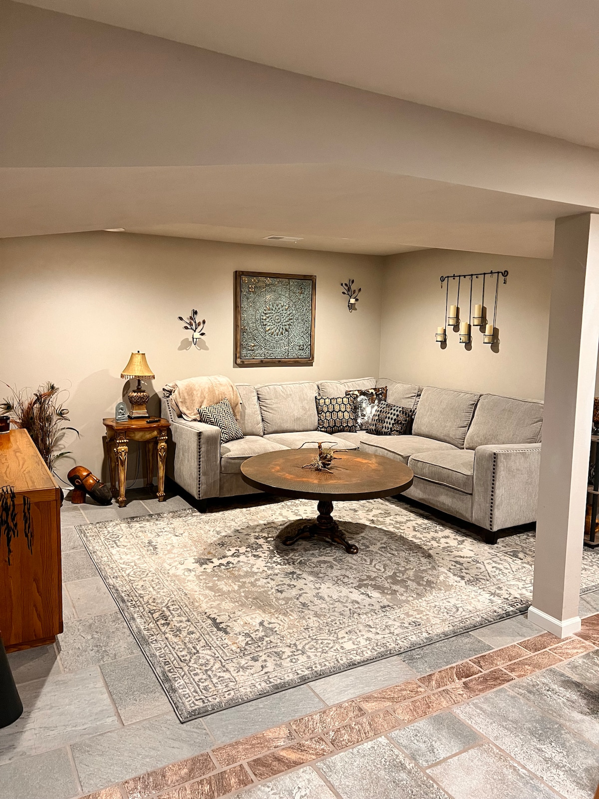 A cozy living area is showcased, featuring a spacious sectional couch and a round wooden coffee table. Soft lighting is provided by a lamp, while decorative wall art and candles add subtle accents. A patterned rug covers the floor, enhancing the inviting atmosphere.