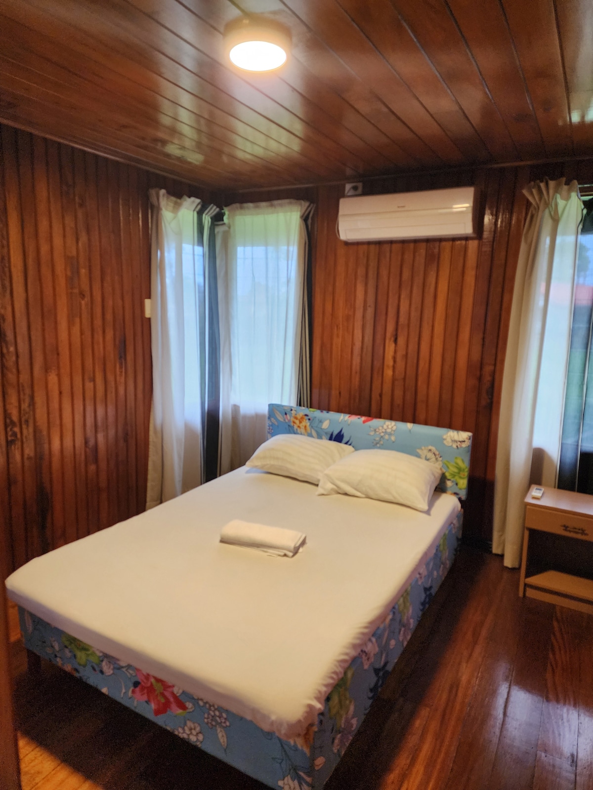 A cozy bedroom features a bed with a floral-patterned base and two soft pillows. Natural light enters through large windows, surrounded by light, flowing curtains. The wood-paneled walls and ceiling create a warm atmosphere, while a small bedside table adds functionality.