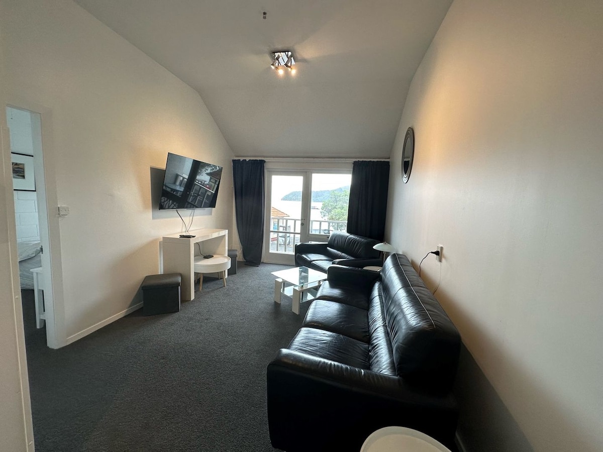 A comfortable living area features a black leather sofa, a coffee table, and a modern TV mounted on the wall. Large windows allow natural light to fill the space and provide views of the surrounding area.