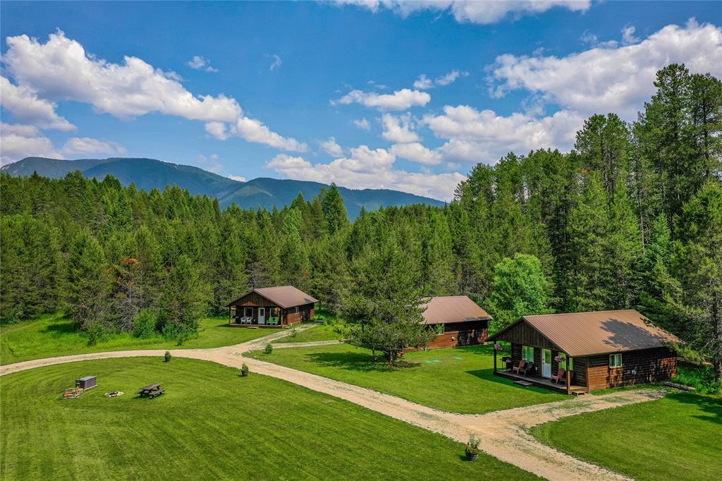 Three rustic cabins are set amidst a lush green landscape, surrounded by tall trees and mountains in the background. A winding gravel path leads to the cabins, with a small firepit area visible on the lawn, creating a serene outdoor space.