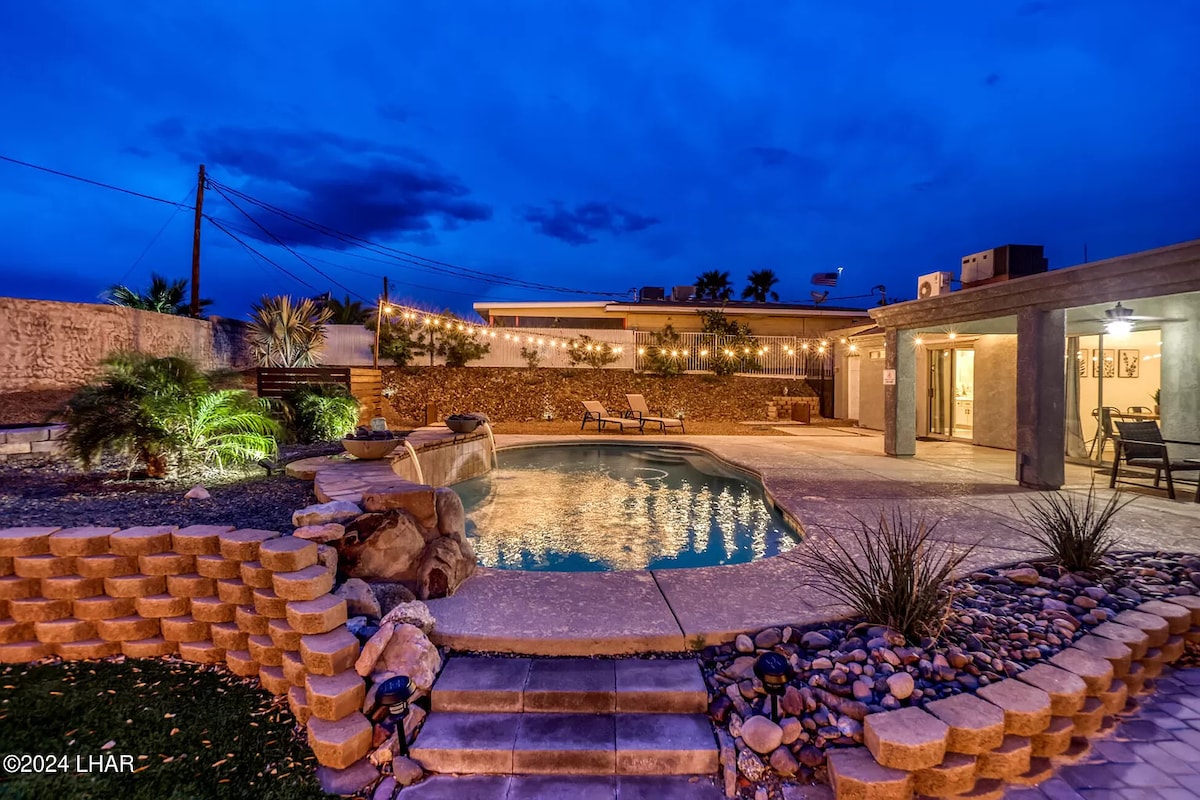 A serene backyard scene is illuminated by soft lighting, showcasing a clear pool reflecting the evening sky. Comfortable lounge chairs are positioned nearby, surrounded by various plants and decorative stones, while the spacious patio area offers ample room for relaxation and outdoor gatherings.