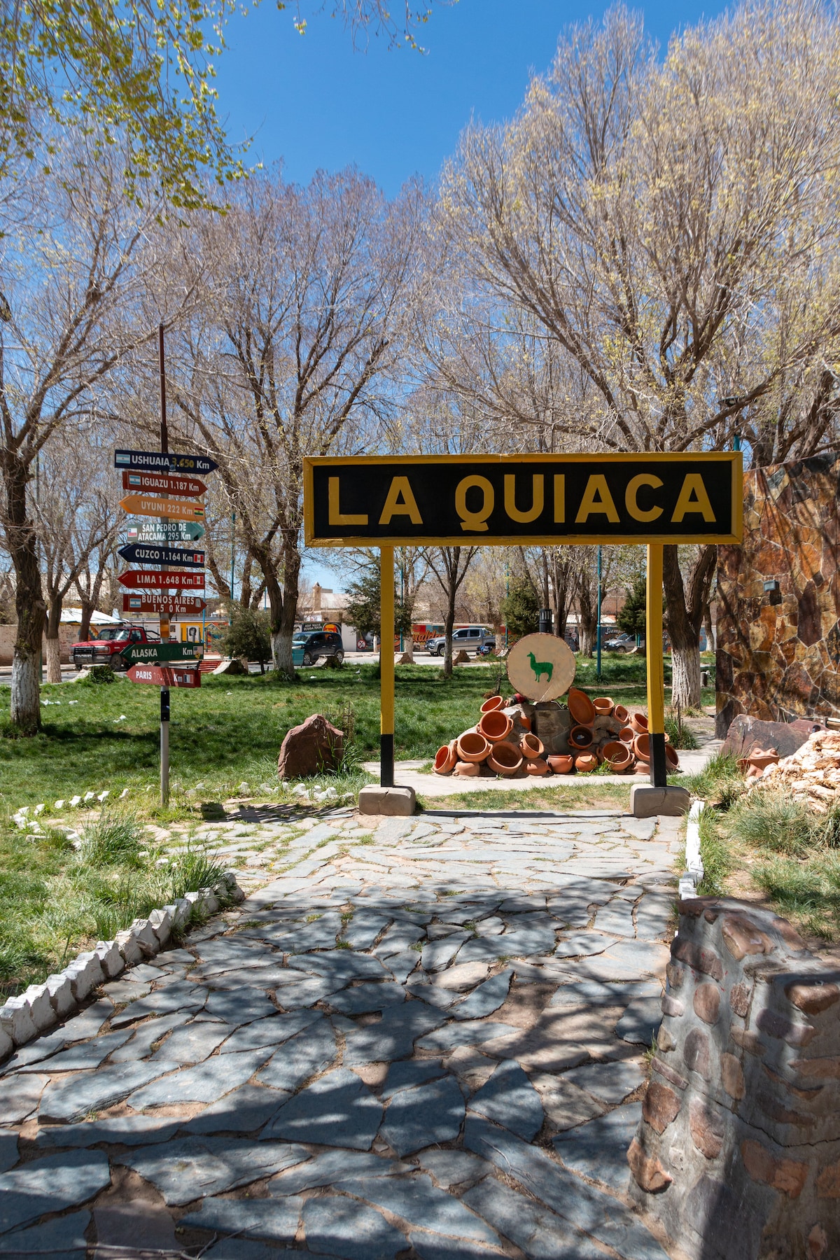 A welcoming sign for 'La Quiaca' is prominently displayed at the entrance. A stone pathway leads through a green area, bordered by trees. Decorative pottery is arranged artistically nearby, creating an inviting environment. Signposts can be seen in the background, indicating various directions.