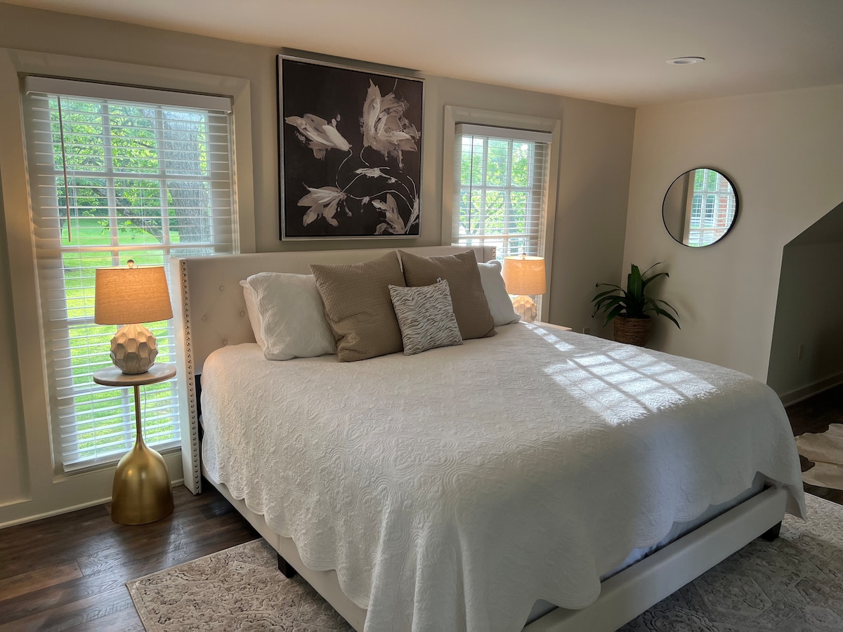A bright sleeping area features a spacious bed dressed in a white quilt. Two bedside lamps provide soft lighting, and large windows invite natural light, revealing a view of the greenery outside. A round mirror and a potted plant add to the serene atmosphere.