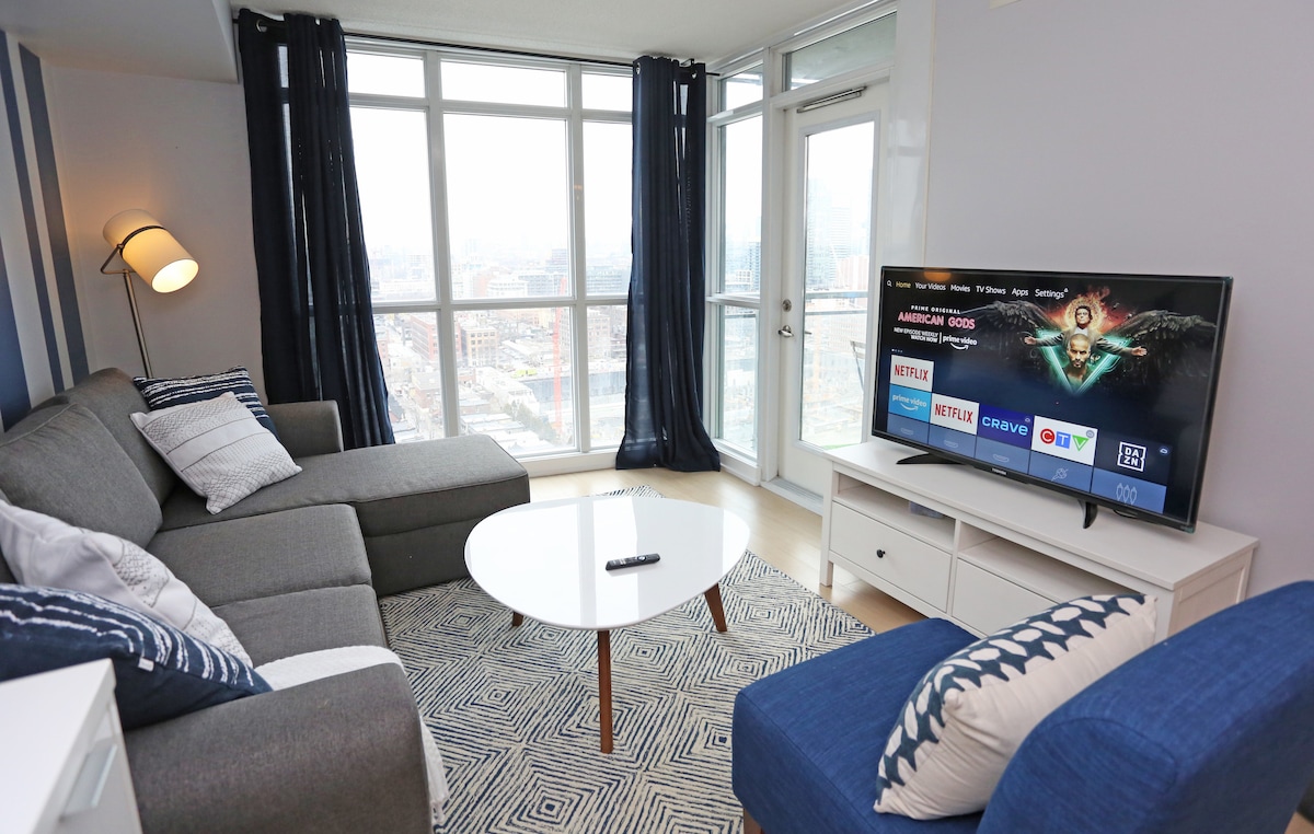 A cozy living area features a gray sectional sofa adorned with decorative pillows. A round coffee table sits in front, positioned on a patterned rug. Large windows provide natural light and views of the city, with sheer dark curtains on either side.