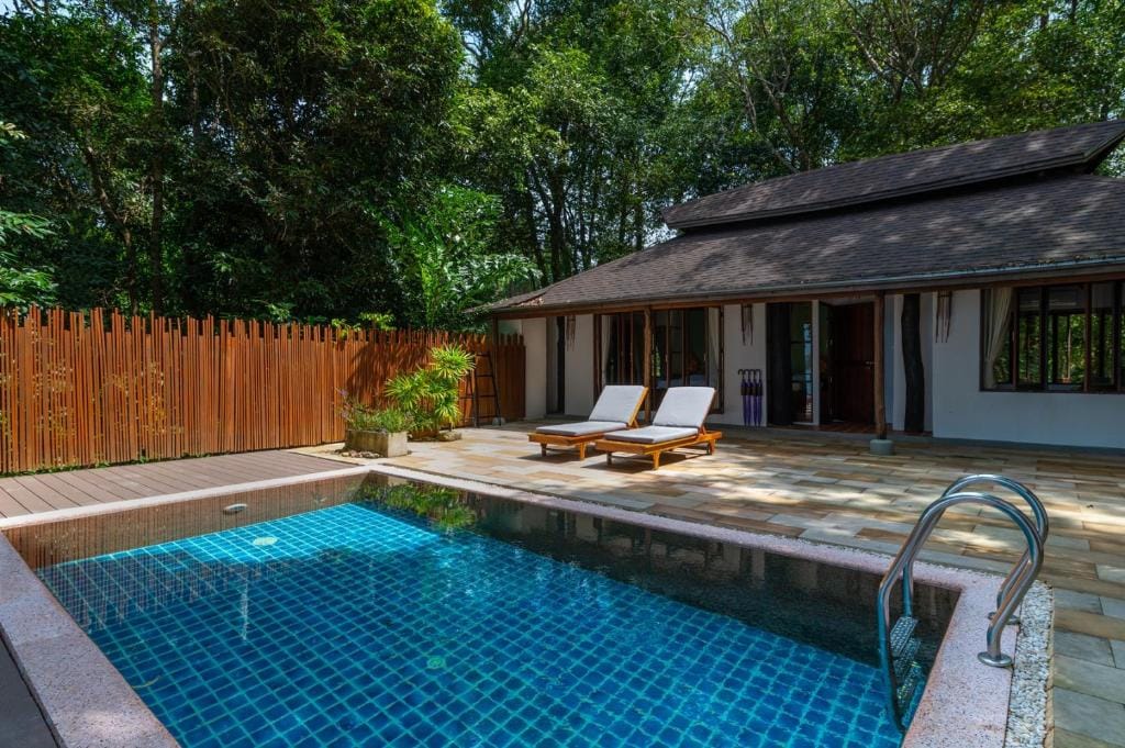 A private outdoor area features a turquoise swimming pool, complemented by two lounge chairs, positioned on a stone patio. Lush greenery surrounds the space, providing a tranquil atmosphere, while a wooden fence offers privacy and natural warmth to the setting.