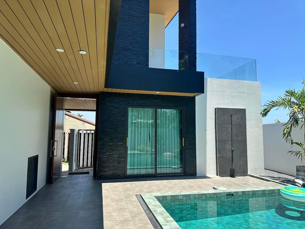 A modern outdoor space features a private pool surrounded by sleek tiles. Floor-to-ceiling glass doors create a seamless transition to the interior. The upper level can be seen with a balcony overlooking the area, brightened by natural light.