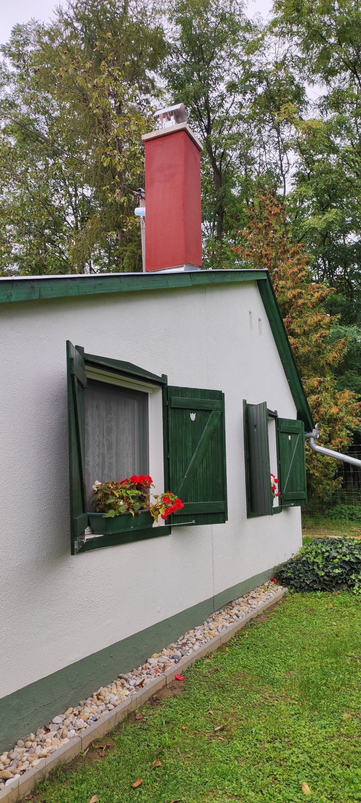 A small house is featured, set against a backdrop of trees. The exterior is painted in soft white with green shutters adorning the windows, each enhanced with vibrant flower pots. A red chimney rises from the roof, complemented by neatly arranged stones along the base.
