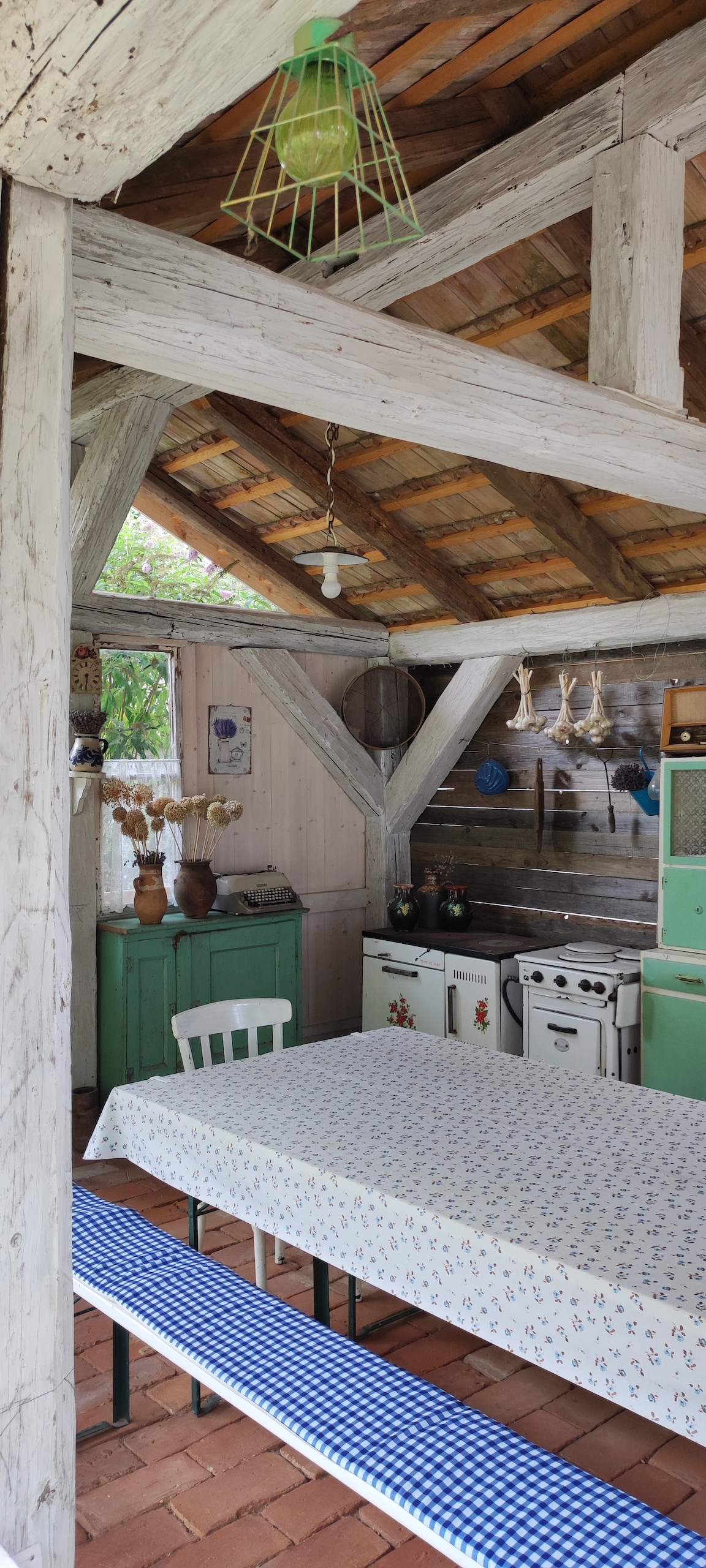 An inviting kitchen area features wooden beams and a light-colored ceiling. A large table is covered with a patterned tablecloth, surrounded by a bench and chairs. Vintage appliances and colorful cabinetry add character, while natural light filters through the windows, enhancing the warm ambiance.