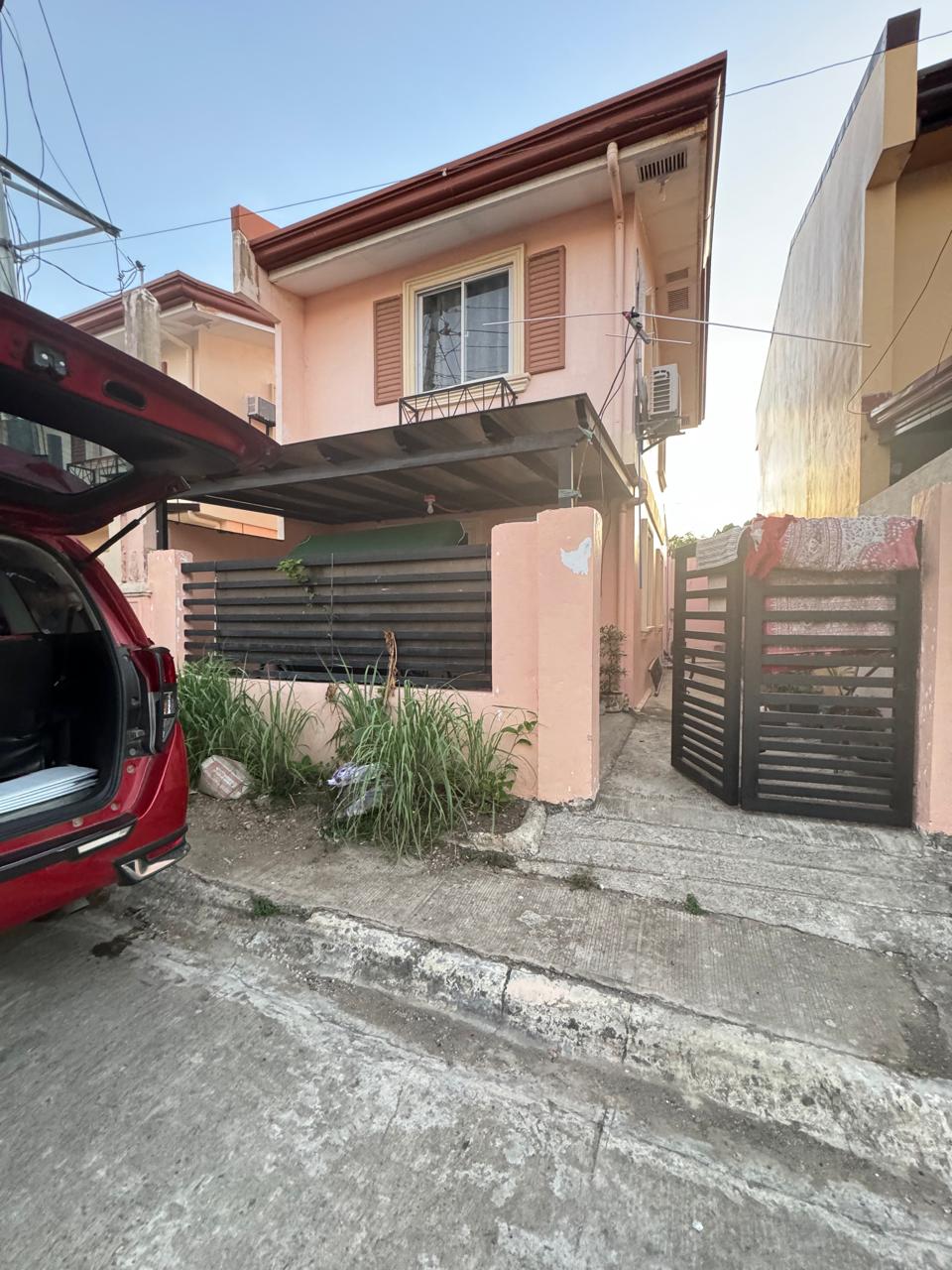 A newly constructed two-story townhouse is visible from the street, featuring a peach-colored exterior and decorative shutters. A covered entry porch is supported by dark pillars, while a black gate leads to the pathway. Lush grass and plants are found near the base of the structure.