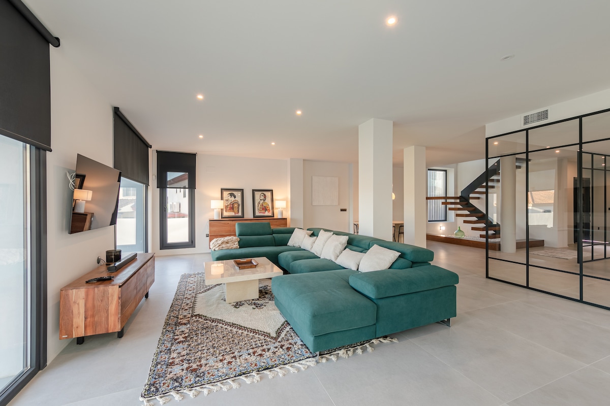 An open living area features a large sectional sofa in a soothing green hue, complemented by a decorative area rug. A modern coffee table sits in front, while natural light pours in through expansive windows. A staircase can be seen in the background, adding depth to the space.