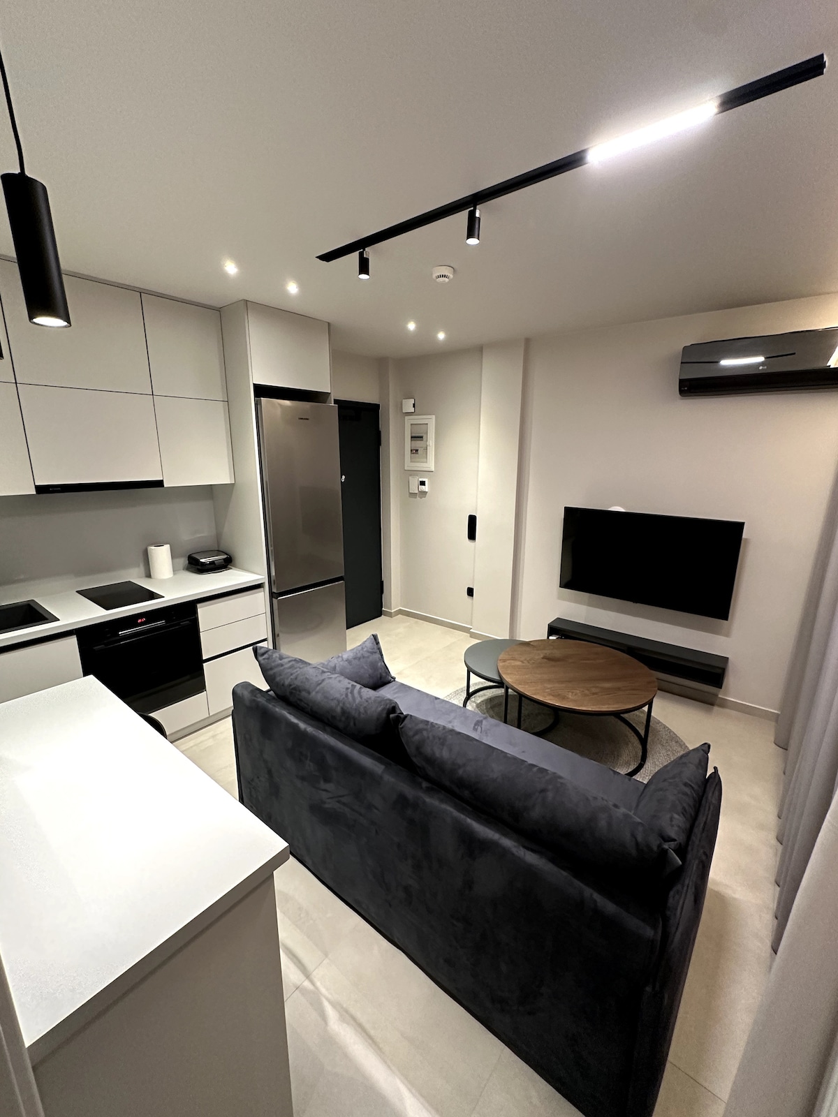 A modern living area showcases a plush gray sofa positioned across from a mounted television. A round coffee table rests in the center, accompanied by sleek kitchen cabinetry in the background. Soft lighting highlights the minimalist decor and spacious layout.