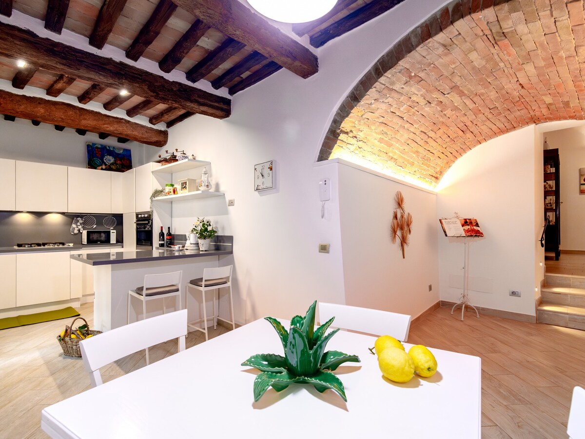 The kitchen and dining area feature exposed wooden beams and a brick archway. A modern dining table with white chairs is accompanied by a decorative green plant and yellow lemons. Sleek kitchen cabinets and an open shelving unit are visible, enhancing the space's functionality.