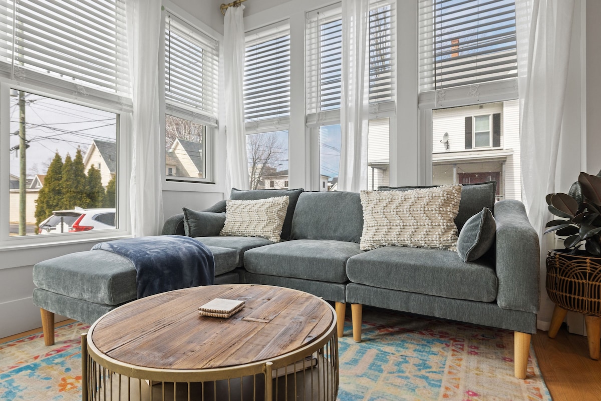 A spacious living area is presented with a comfortable sectional sofa in a soft grey fabric. A wooden coffee table sits at the center, complementing the vibrant area rug. Natural light floods in through large windows adorned with sheer curtains, providing a bright atmosphere.