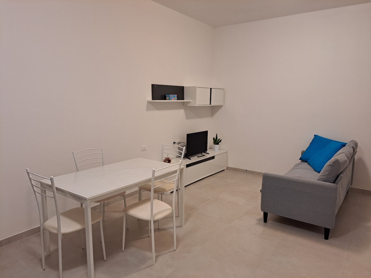 A modern living space features a dining table with four chairs, positioned adjacent to a wall-mounted television. A light grey sofa with a blue cushion is placed against the wall, complementing the neutral color palette of the room. The flooring is tiled and light-toned.