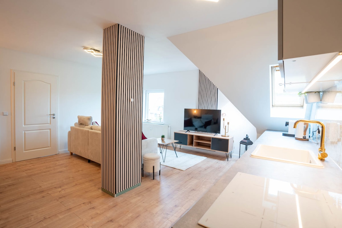 A modern living area features sleek wooden flooring, with a comfortable couch and a small armchair positioned near a wall-mounted television. Open shelving displays decorative items, while large windows let in natural light, complementing the neutral color palette of the space.