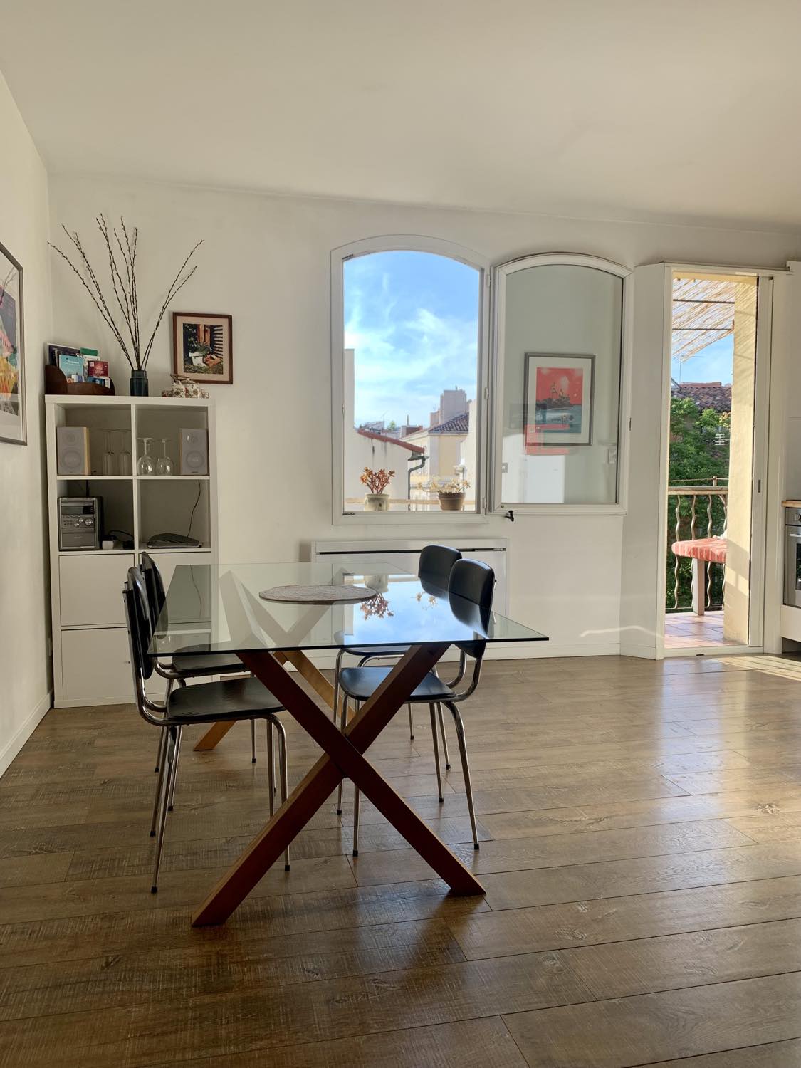 A modern dining area features a glass table surrounded by four black chairs. Natural light floods in through large windows, complementing the light-colored walls and wooden flooring. The space also includes decorative artwork and a small cabinet, enhancing its functionality.