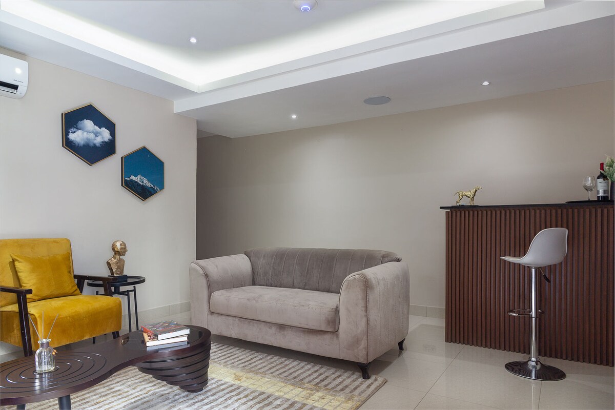 A modern living area features a plush sofa in neutral tones complemented by a yellow accent chair. A sleek bar counter, paired with a minimalist stool, is situated against a wall. Wall art adds character, while soft lighting creates an inviting environment.