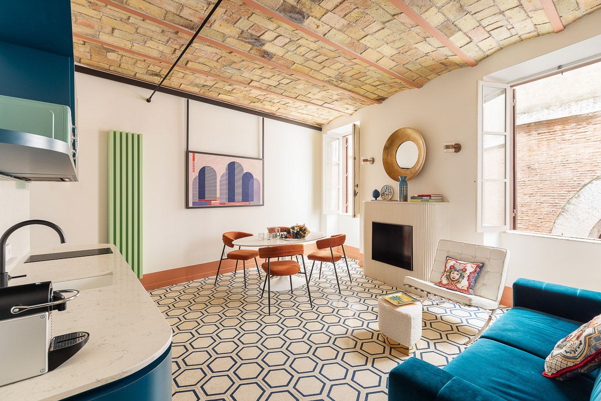 An inviting living space showcases a vibrant blue sofa and a circular mirror above a modern fireplace. A stylish dining table with wooden chairs is set against a backdrop of textured brick ceilings and large windows, allowing natural light to fill the room.