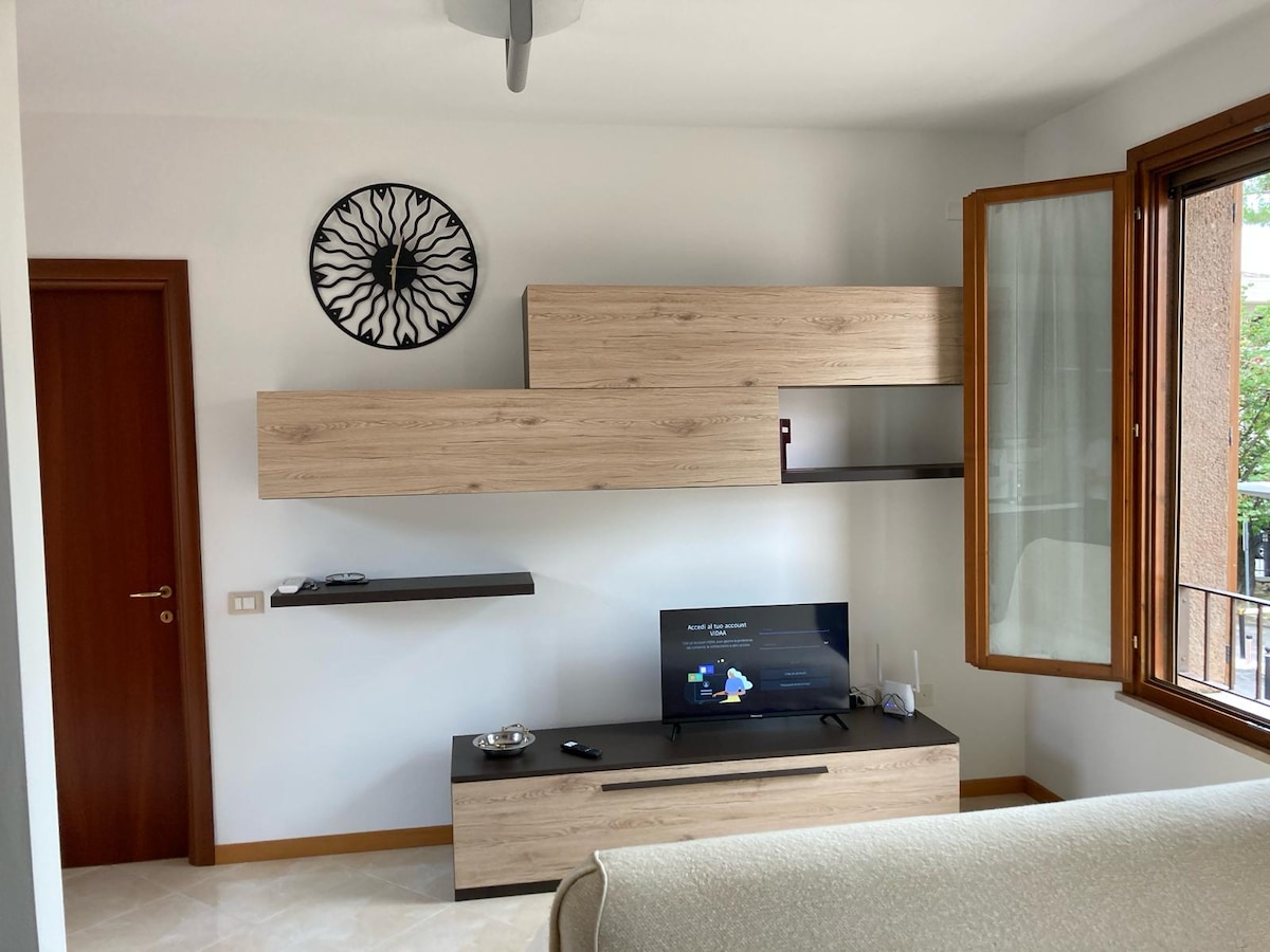 A bright living area features a modern wooden entertainment unit with a television and decorative wall shelf. Natural light enters through a window adorned with light curtains, while a circular wall art piece adds a touch of design.