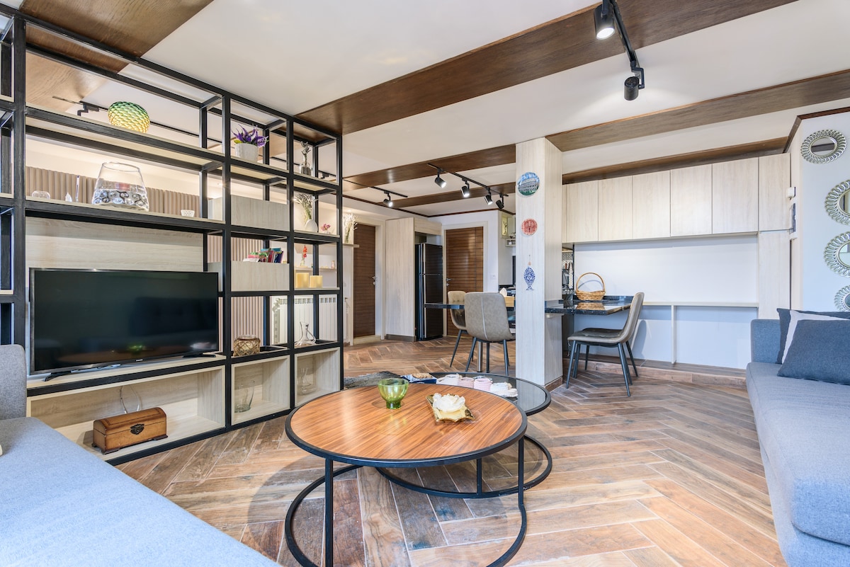 The living space features an open layout with a stylish shelving unit and a comfortable seating area. A round coffee table is positioned at the center, surrounded by gray sofas. Natural light fills the room, highlighting the wooden flooring and contemporary decor.