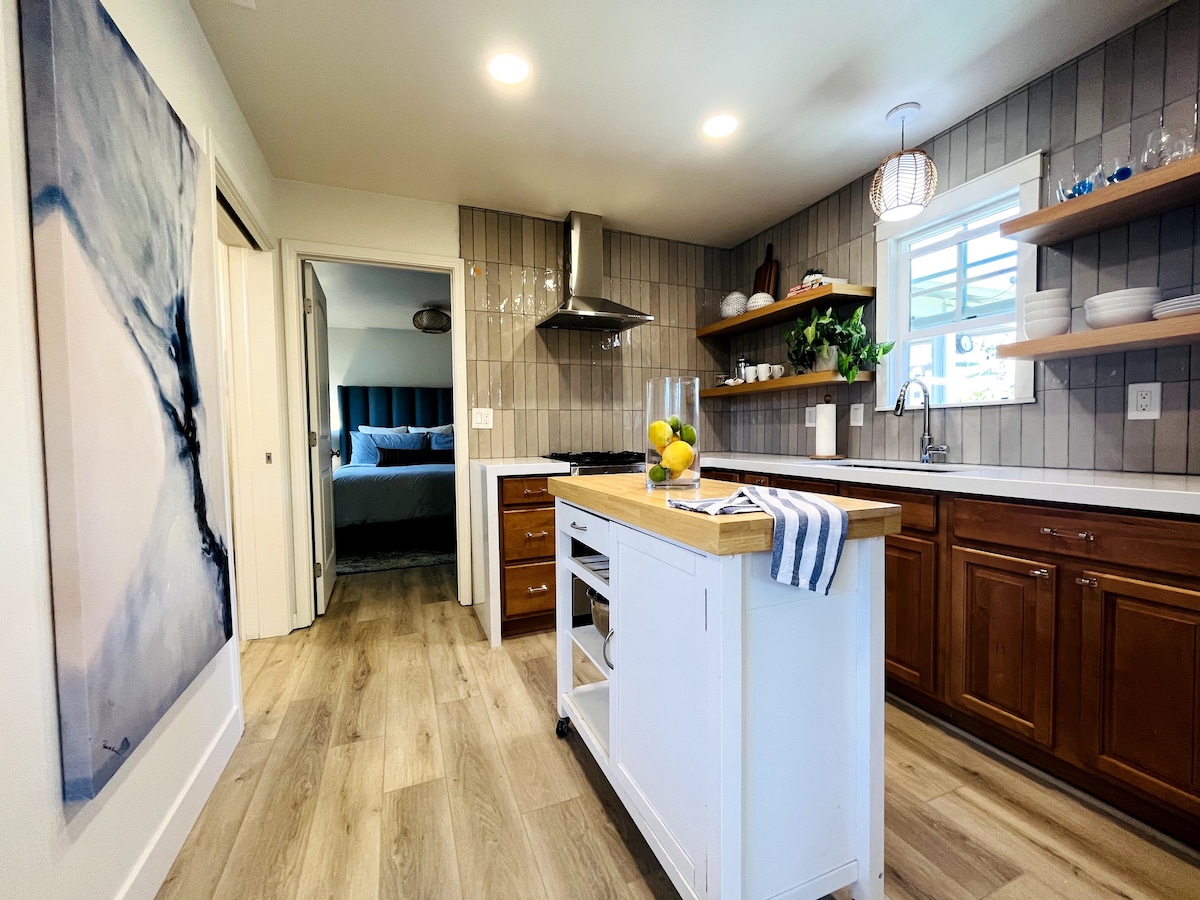 A modern kitchen features light wood cabinetry and open shelving. An island with a striped towel holds fresh fruit. Stainless steel appliances complement the neutral-toned walls, and natural light floods the space from a nearby window. The bedroom area is visible through an open door.