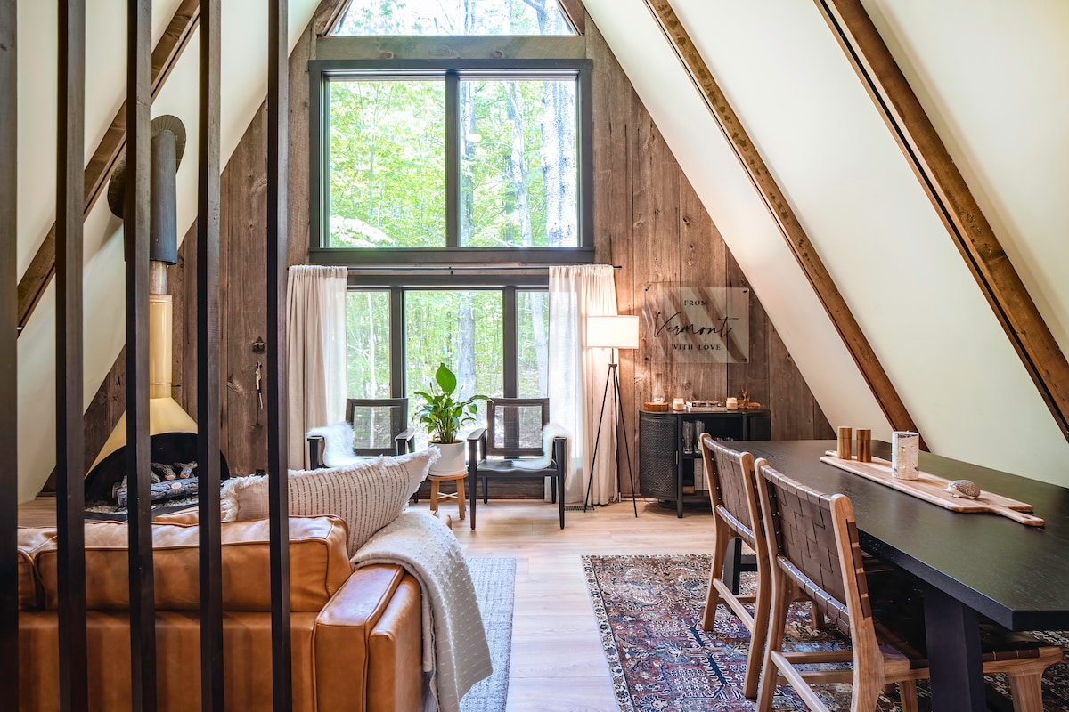 The inviting living area features high A-frame ceilings and large windows allowing natural light to fill the space. A comfortable leather sofa and armchairs are arranged around a decorative area rug. A dining table is positioned beside the fireplace, enhancing the cozy atmosphere.